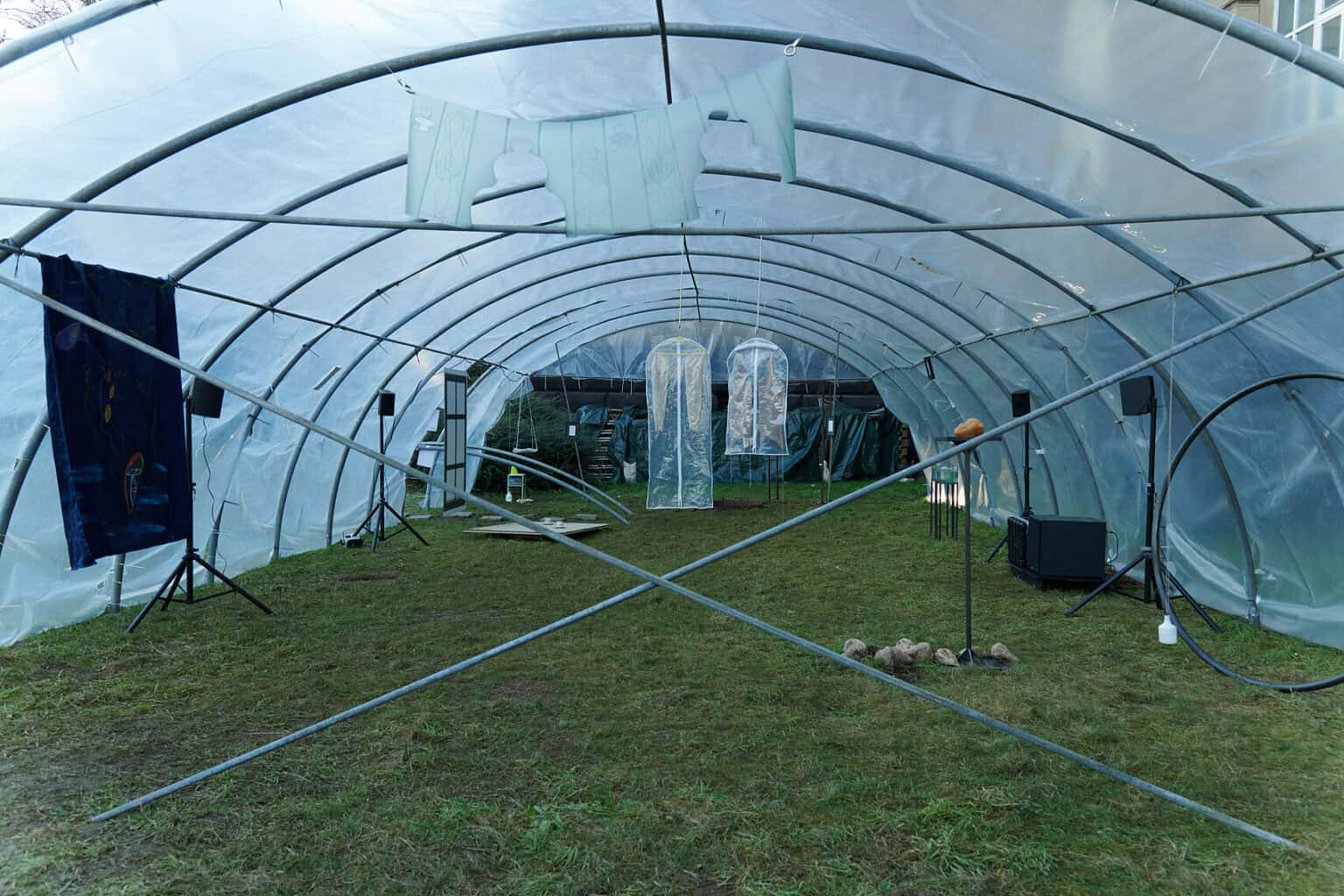 Foto: Ansicht eines mit transparenter Folie abgehangenen, tunnelartigen Metallkonstruktion, im Außenbereich, auf Rasen. Innerhalb der Konstruktion sind Metallstangen in Kreuzform angebracht. Von den Metallstrebender der Konstruktion hängen Objekte und auf dem Boden stehen weitere Objekte.
