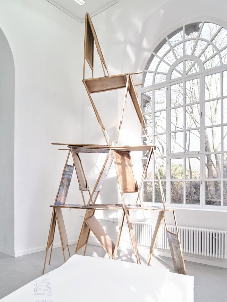 Foto: Ansicht eines Raumes mit weißen Wänden, hellgrauem Boden und einem großen Bogenfenster. Im Raum befindet sich hohes Objekt, das aus gestapelten Gegenständen besteht.