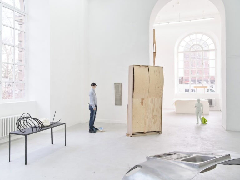 Foto: Aufnahme eines Raumes mit weißen Wänden, hellgrauem Boden und zwei großen Bogenfenstern. An der Wand montiert und frei stehend im Raum befinden sich Objekte aus unterschiedlichem Material.