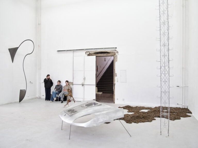 Foto: Aufnahme eines Raumes mit weißen Wänden, hellgrauem Boden und einer Tür. Im Raum befinden sich Objekte in unterschiedlichen Größen und aus unterschiedlichen Materialien. Sie stehen frei im Raum, lehnen an der Wand oder liegen auf dem Boden. Im Raum befinden sich außerdem drei Menschen, die stehen und auf Stühlen sitzen.