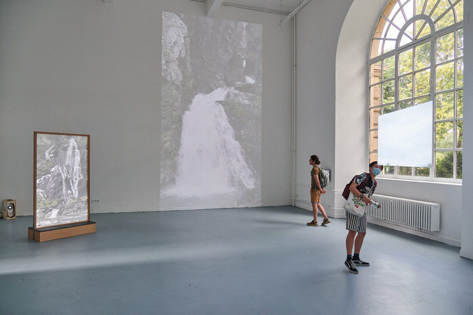 Foto: Ein Raum mit weißen Wänden und grauem Boden. Auf eine der Wand wird eine große, quadratische Projektion geworfen. im Raum steht ein Objekt, dass von einer Projektion angestrahlt wird. An einer anderen Wand befindet sich ein kleiner, eingeschalteter Bildschirm. Von der Decke hängt eine Platte, die von einer Projektion angestrahlt wird. Im Raum befinden sich zwei Personen.