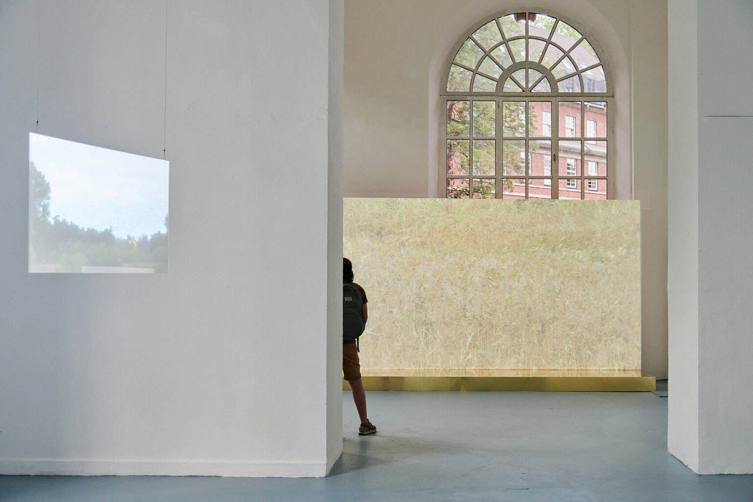 Foto: Ein Raum mit weißen Wänden, grauem Boden und einem Fenster. Im Raum befindet sich ein großes, flächiges Objekt, dass beinahe so breit ist wie der Raum. Es verdeckt Teile des Fensters. Von der Decke hängt eine Platte, die von einer Projektion angestrahlt wird. Im Raum befindet sich ein Mensch.