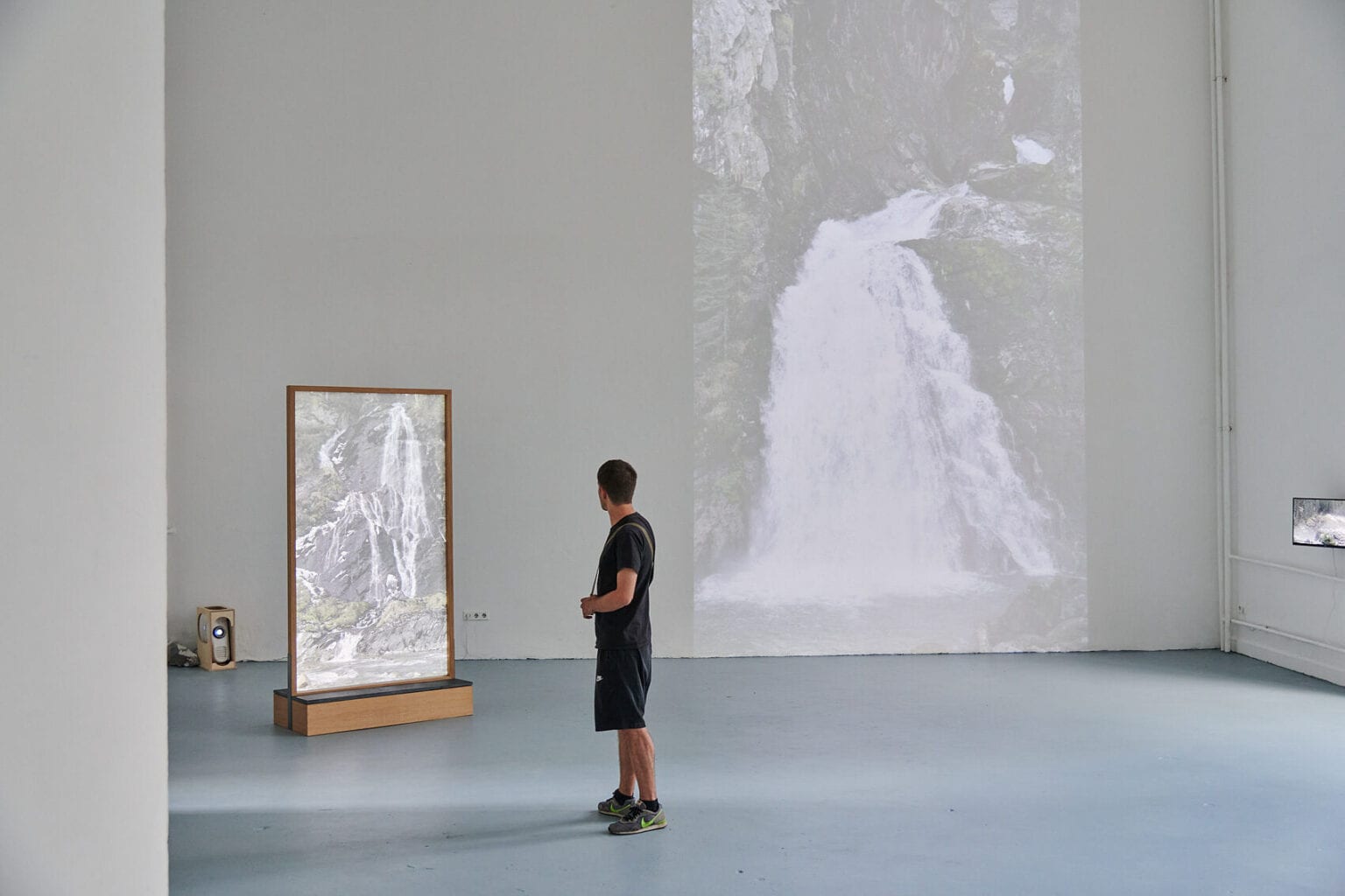 Foto: Ein Raum mit weißen Wänden und grauem Boden. Auf eine der Wand wird eine große, quadratische Projektion geworfen. im Raum steht ein Objekt, dass von einer Projektion angestrahlt wird. An einer anderen Wand befindet sich ein kleiner, eingeschalteter Bildschirm. Im Raum befindet sich eine Person.