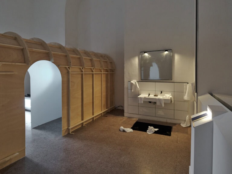 Foto: Ansicht eines Raumes mir weißen Wänden und einem rötlichen Fliesenboden. Um Raum befinden sich verschiedene Objekte und Konstruktionen, darunter ein Waschbecken das von weißen Fliesen umgeben ist, ein Spiegel mit zwei Leuchten, herumliegende weiße Handtücher, ein schwarzer Badvorleger, ein an die Wand gebautes Pult, auf dem ein Buch liegt, das von einer Lampe angeleuchtet wird. Links im Bild befindet sich eine Konstruktion die eine bogenförmige Türaussparung hat.