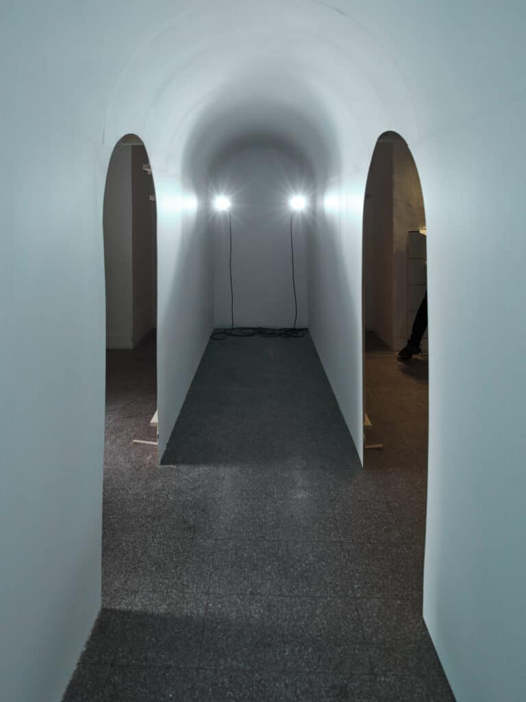 Foto: Ansicht eines schmalen, weißen Gangs mit zwei bogenförmigen Türaussparungen. Im Gang sind zwei Lichter installiert, die an Kabeln hängen.