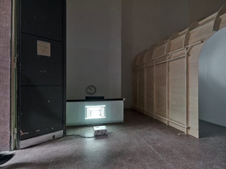 Foto: Ansicht eines abgedunkelten Raumes mit weißen Wänden und rötlichem Fliesenboden. Eine schwarze geöffnete Flügeltür ist zu sehen. Im Raum befinden sich verschiedene Objekte und Konstruktionen.