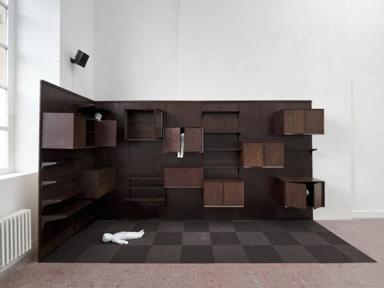 oto: Ansicht eines Raumes mit weißen Wänden, rötlichem Fliesenboden und einem Fenster. Links im Bild befindet sich eine verschachtelte Schrank- und Regalkonstruktion aus unterschiedlichen Materialien mit vielen dunklem Elementen. In ihr liegen zwei helle Objekte. Der Boden in diesem Teil des Raumes ist mit einem hell- und dunkelbraunen Schachbrettmuster ausgekleidet.