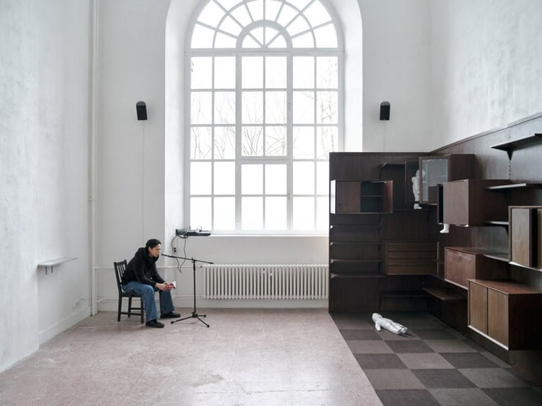 Foto: Ansicht eines Raumes mit weißen Wänden, rötlichem Fliesenboden und einem großen Bogenfenster. Links im Raum sitzt eine Person gebeugt auf einem Stuhl. Vor ihr steht ein Mikrofonständer. Links an der Wand ist ein weißes Regal montiert. Über dem Fenster hängt ein Lautsprecher. Rechts im Raum befindet sich eine verschachtelte Schrank- und Regalkonstruktion aus unterschiedlichen Materialien mit vielen dunklem Elementen. Der Boden in diesem Teil des Raumes ist mit einem hell- und dunkelbraunen Schachbrettmuster ausgekleidet.