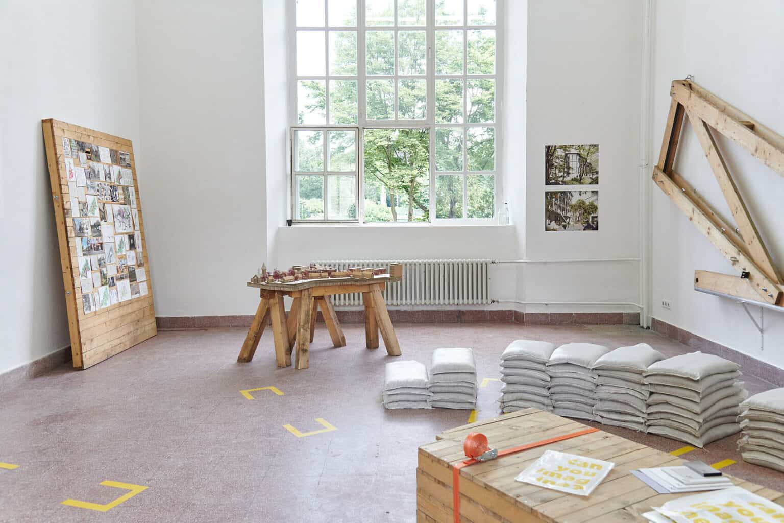 Foto: Ein Raum mit weißen Wänden, rotem Steinboden und einem großen Fenster. An den Wänden des Raumes befindet sich eine große Konstruktion aus Holzlatten und Bilder im Mittelformat. Auf dem Boden des Raumes liegen Stapel aus gefüllten Säcken, eine Tischkonstruktion auf Holz, auf der sich Objekte befinden und zusammengeschnürte, aufeinander gestapelte Paletten. Auf den Paletten liegen Objekte. An die Wand gelehnt steht eine Tafel aus Holzlatten, auf der kleinformatige, bemalte Papiere angebracht sind.