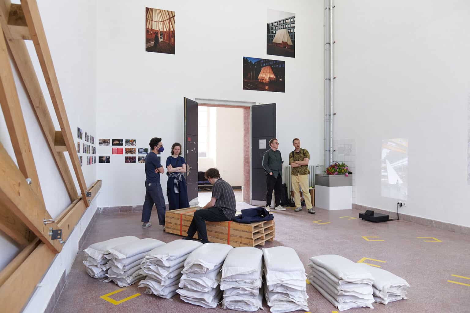 Foto: Ein Raum mit weißen Wänden und rotem Steinboden. An den Wänden des Raumes befindet sich eine große Konstruktion aus Holzlatten und Bilder in Klein- und Mittelformaten, auf unterschiedlicher Höhe in Gruppen hängend. Auf dem Boden des Raumes liegen Stapel aus gefüllten Säcken, zusammengeschnürte, aufeinander gestapelte Paletten und ein Beamer, der eine Projektion an die Wand wirft. Im Raum stehen und Sitzzen mehrere Menschen.