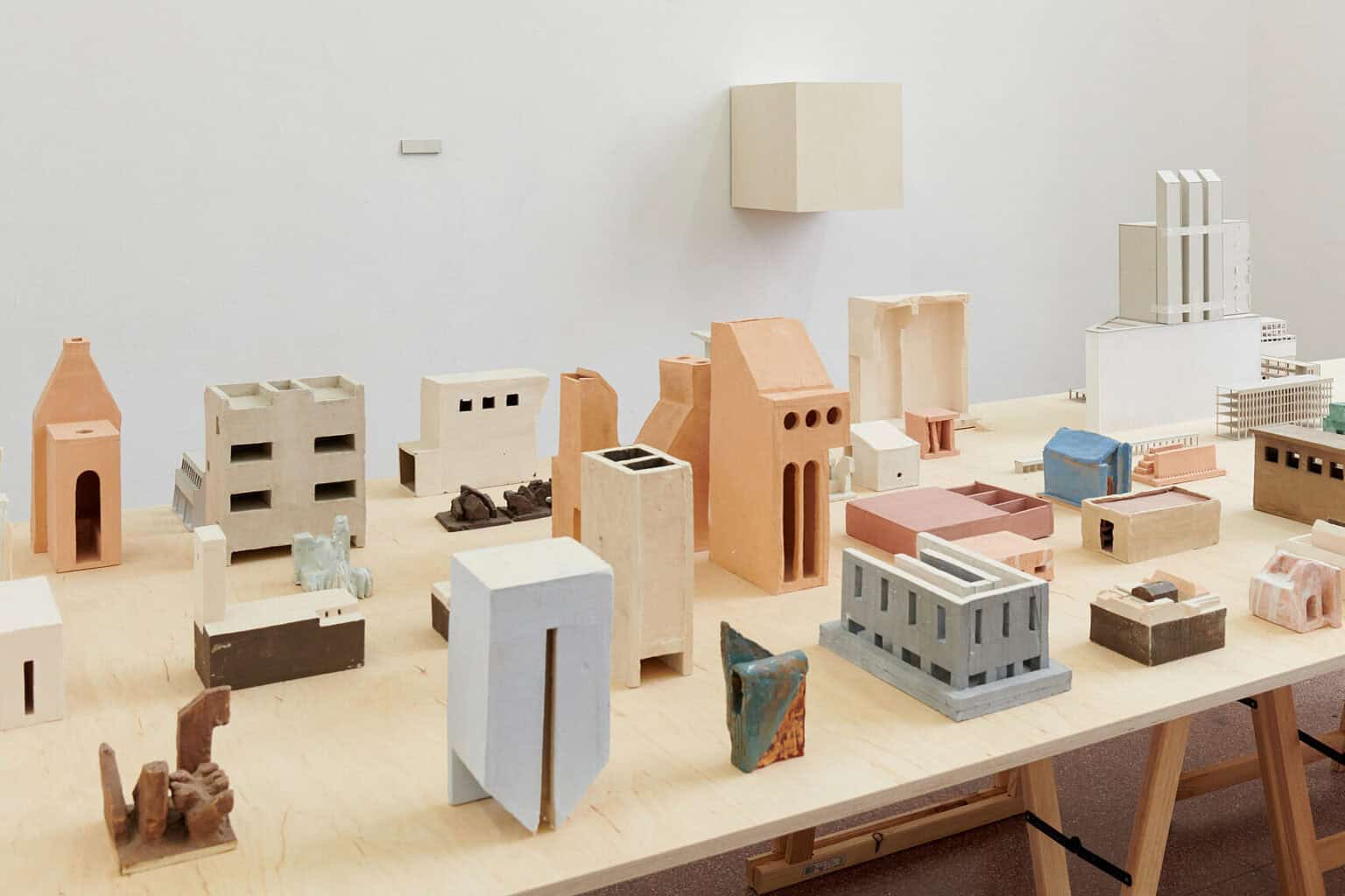 Foto: Ansicht eines Raumes mit weißen Wänden. Im Raum befindet sich eine Holzplatte auf Böcken, auf der viele Objekte stehen. An der Wand hängt ein weiteres Objekt.