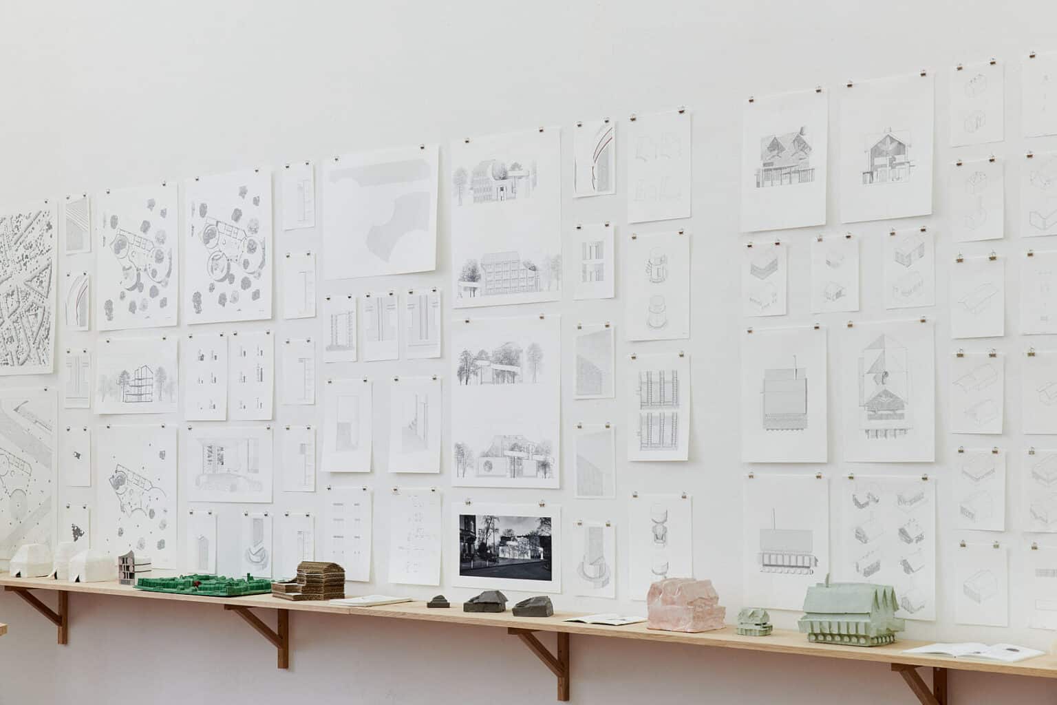 Foto: Ansicht einer Wand mit weißen Wänden. Unten an der Wand befindet sich eine Reihe aus an der Wand montierten Regalbrettern, auf der Objekte, Hefte und Blätter liegen. Oberhalb des Regals ist eine große Menge von Papierarbeiten in mehreren Reihen über- und nebeneinander an der Wand befestigt.