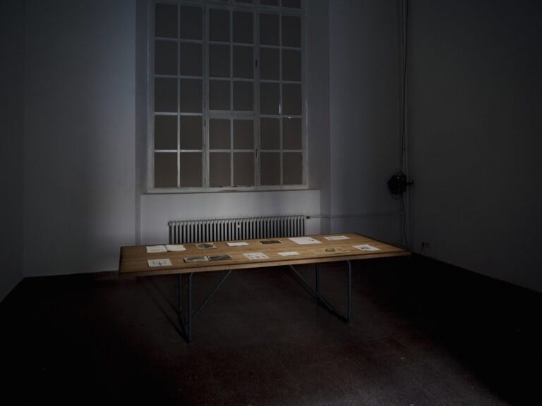 Foto: Ansicht eines abgedunkelten Raumes mit weißen Wänden, dunklem Boden und einem großen Fenster. Im Raum steht ein Tisch, der angeleuchtet wird. Auf dem Tisch liegen Papiere.