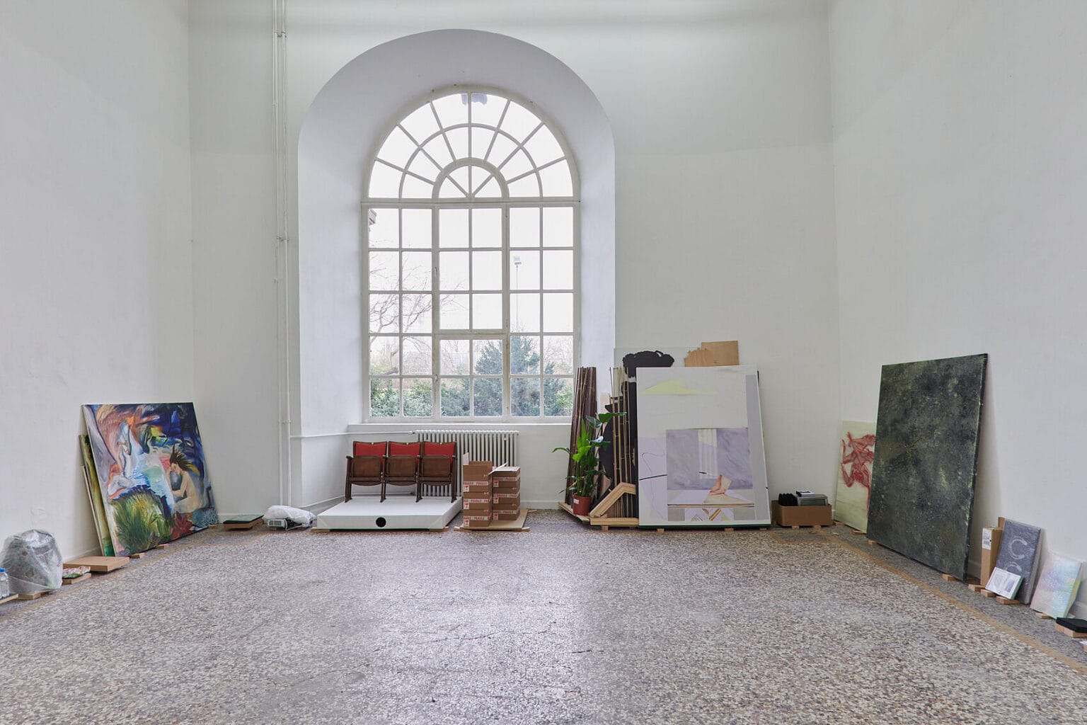 Foto: Ansicht eines Raumes mit weißen Wänden, mehrfarbigem Steinboden und großem Bogenfenster. Vor den Wänden stehen auf dem Boden verschiedenartige Objekte und Möbel.