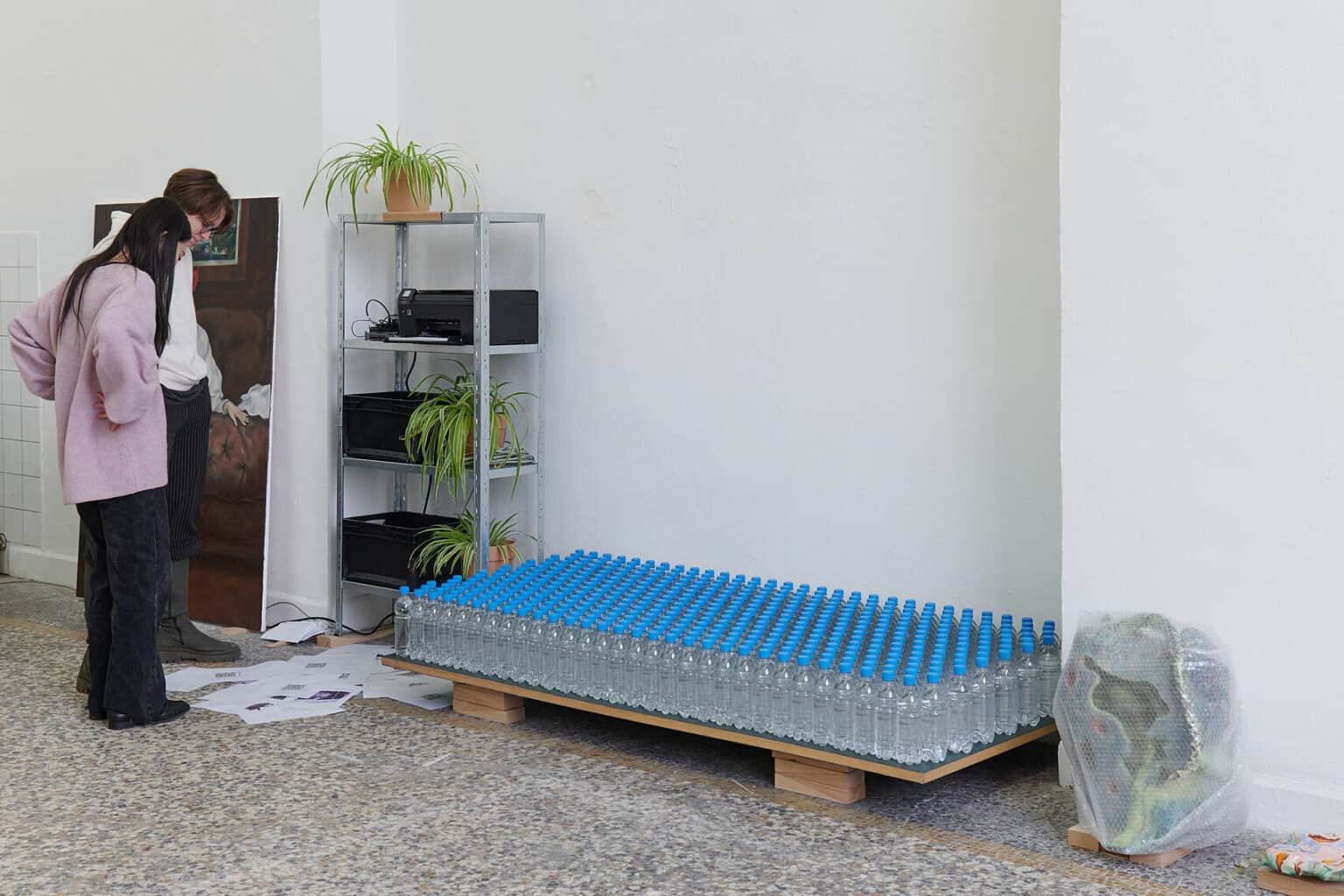 Foto: Ansicht eines Raumes mit weißen Wänden und mehrfarbigem Steinboden. Vor der Wand stehen auf dem Boden verschiedenartige Objekte und Möbel. Zwei Menschen stehen im Raum vor den Dingen.