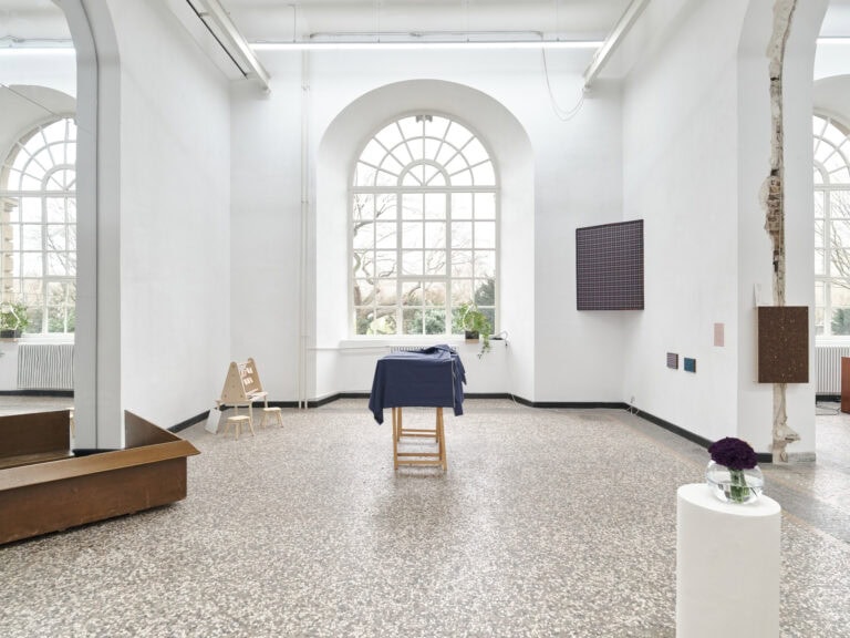 Foto: Ansicht eines Raumes mit weißen Wänden, schwarzer Fußleiste, mehrfarbigen Steinboden und einem großen Bogenfenster. In eine bogenförmigen, wandhohen Aussparung ist eine spiegelnde Fläche eingelassen. Vor der Spiegelfäche steht eine braunfarbige Konstruktion am Boden. Im Raum und an den Wänden befinden sich Objekte. Manche von Ihnen stehen auf dem Boden, andere auf Sockeln.