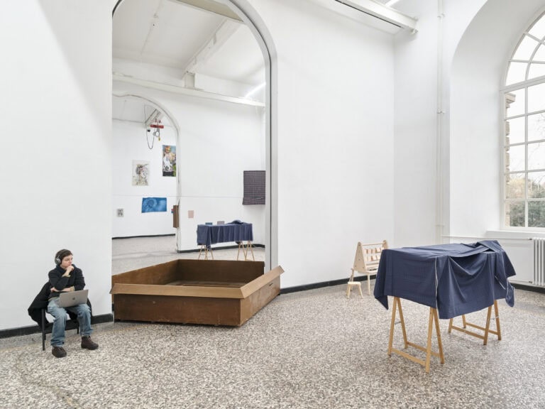 Foto: Ansicht eines Raumes mit weißen Wänden, schwarzer Fußleiste, mehrfarbigen Steinboden und einem großen Bogenfenster. In eine bogenförmigen, wandhohen Aussparung ist eine spiegelnde Fläche eingelassen. Darin spiegelt sich der Rest des Raumes und eine Vielzahl von Objekten, die sich an den Wänden des Raumes befinden. Vor der Spiegelfäche steht eine braunfarbige Konstruktion am Boden. Im Raum befinden sich Skulpturen. Links im Bild sitzt eine Person mit Köpfhören und einem Laptop auf dem Schoß.