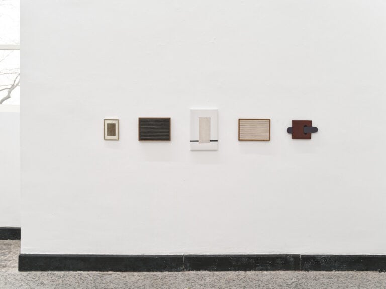 Foto: Ansicht eines Raumes mit weißen Wänden, einem Fenster, einer schwarzen Fußleiste und einem mehrfarbigen Steinboden. An den Wänden hängen auf gleicher Höhe Objekte in einer Reihe.