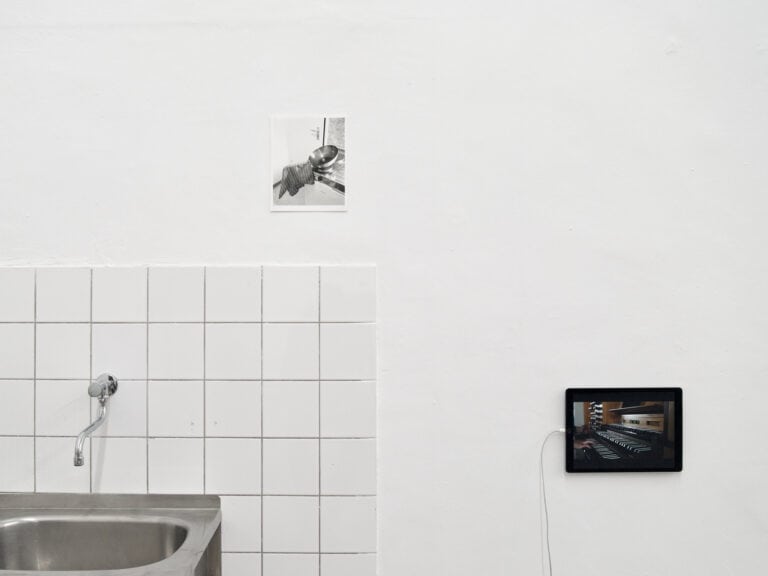 Foto: Nahaufnahme eines Raumes mit weißer Wand und einem Metallwaschbecken, das von weißen Fliesen umgeben ist. An der Wand befindet sich eine Fotografie und ein iPad, das eine Abbildung zeigt.