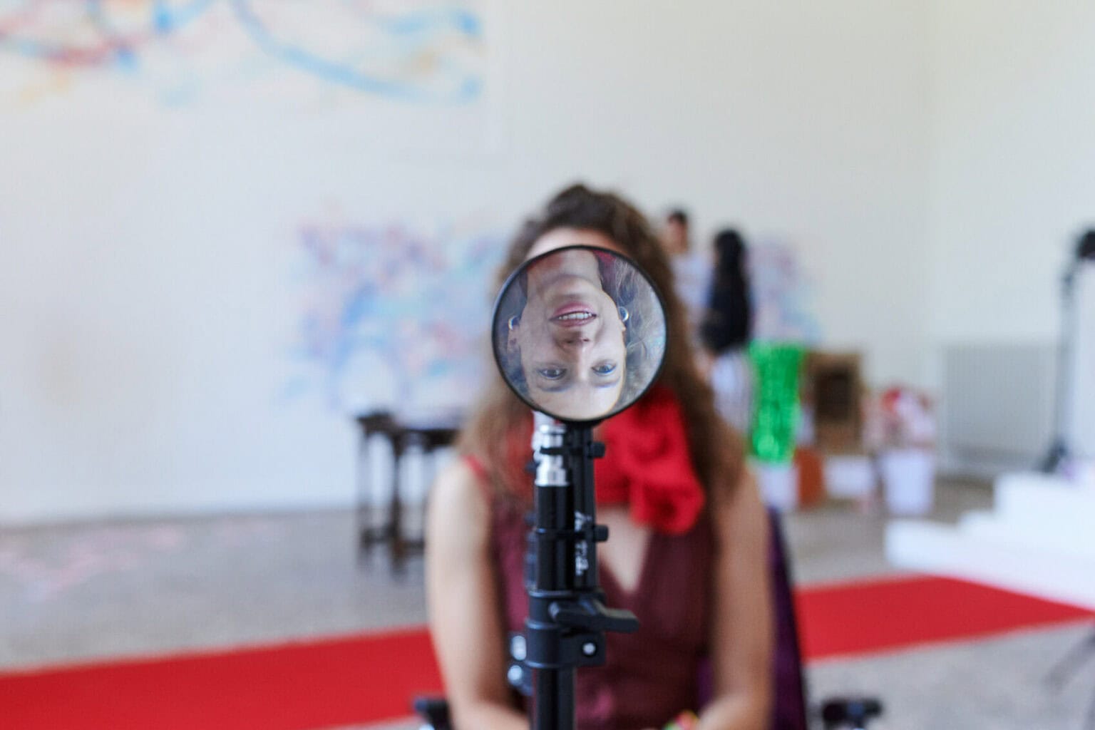 Foto: Eine an einem Stativ befestigte Lupe. Hinter der Lupe ist unscharf eine Person zu erkennen. In der Lupe ist ihr umgedrehtes Gesicht zu erkennen.