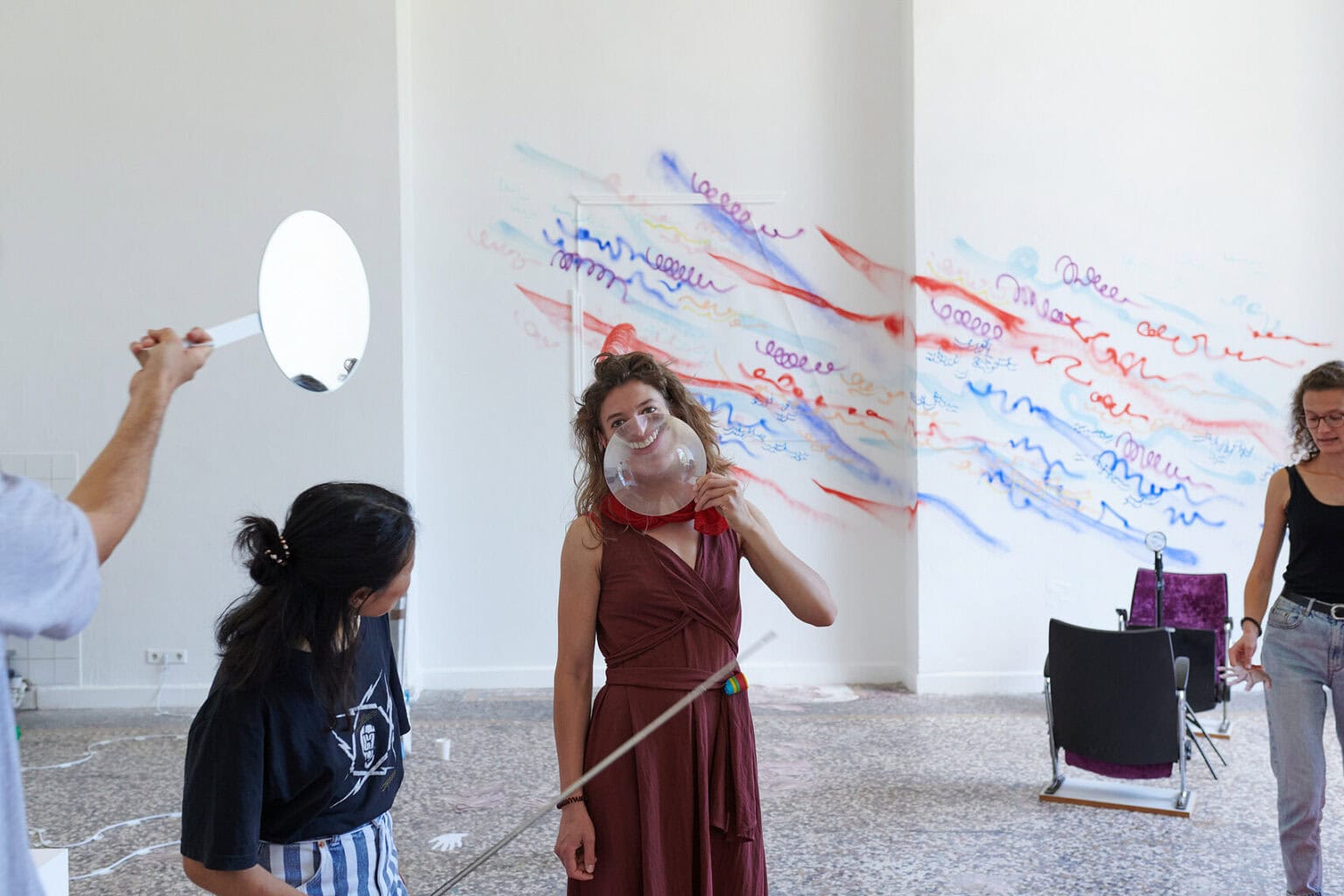 Foto: Ein Raum mit weißen Wänden, mehrfarbigem Steinboden und einem großen Fenste. Teile der Wände sind mit bunten Linien bemalt. Im Raum verteilt befindet sich eine Vielzahl unterschiedlicher Objekte, darunter ein ausgerollter, roter Teppich, Lautsprecher, ein Ventilator, ein Tisch, eine Kleiderstange, eingepackte Leinwände, Pappboxen usw. Durch den Raum bewegen sich zwei Menschen, die mit den Objekten interagieren. Eine der Personen hält sich ein lupenartiges Glas vor ihr Gesicht, dass Teile ihres Gesichtes stark vergrößert.