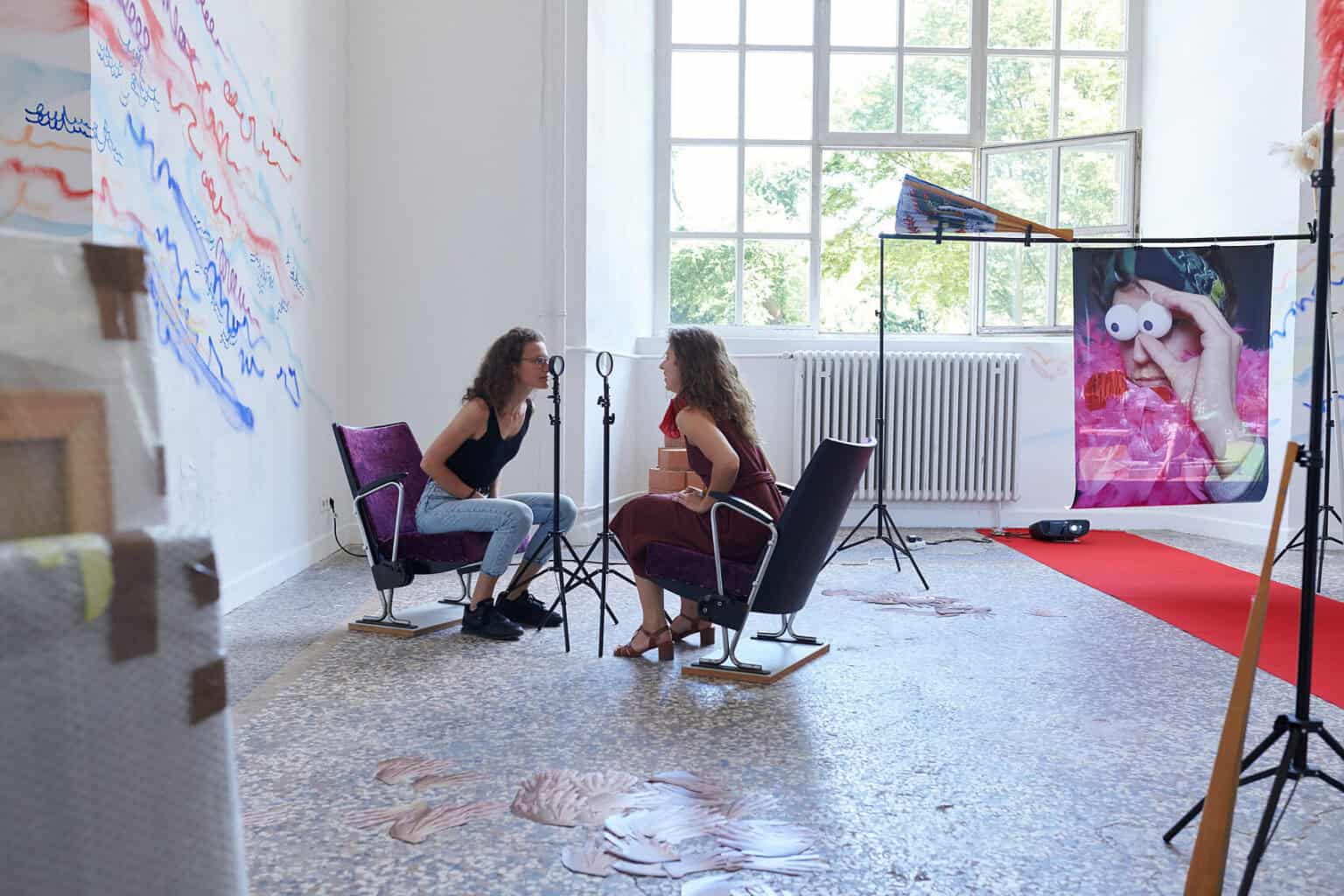 Foto: Ein Raum mit weißen Wänden, mehrfarbigem Steinboden und einem großen Fenste. Teile der Wände sind mit bunten Linien bemalt. Im Raum verteilt befindet sich eine Vielzahl unterschiedlicher Objekte, darunter ein ausgerollter, roter Teppich, Lautsprecher, ein Ventilator, ein Tisch, eine Kleiderstange, eingepackte Leinwände, Pappboxen usw. Durch den Raum bewegen sich zwei Menschen, die mit den Objekten interagieren.