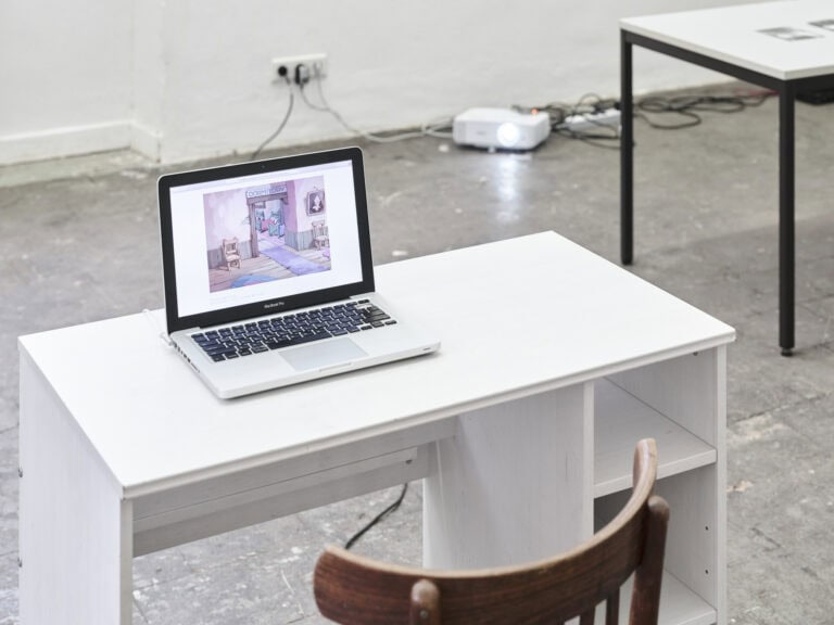 Foto: Nahaufnahme eines Laptops, der auf einem weißen Tisch steht, vor einem grauen, gekachelten Boden. Im Hintergrund steht Technik.