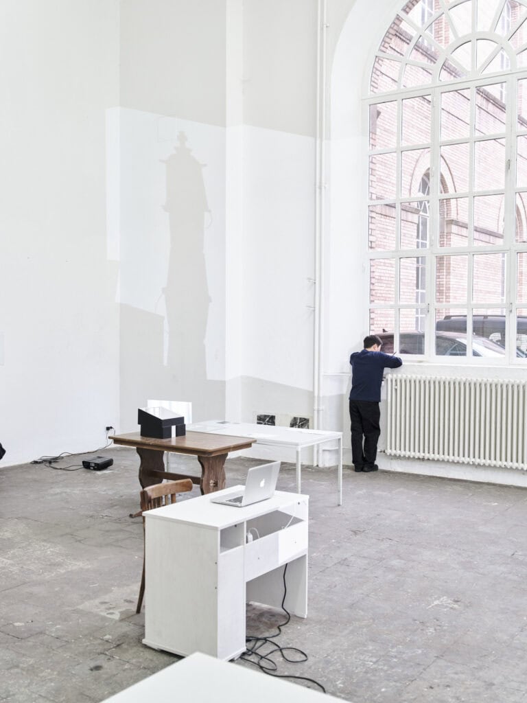 Foto: Ansicht eines Raumes mit weißen Wänden, grauem, gekachelten Boden und einem großen Bogenfenster. Im Raum befinden sich Möbel, darunter Tische und Stühle und weitere Objekte. Manche der Objekte hängen an der Wand, andere liegen oder stehen auf Tischen. Auf einem Teil der Wand ist eine Lichtprojektion geworfen. Eine Person steht am Fenster.