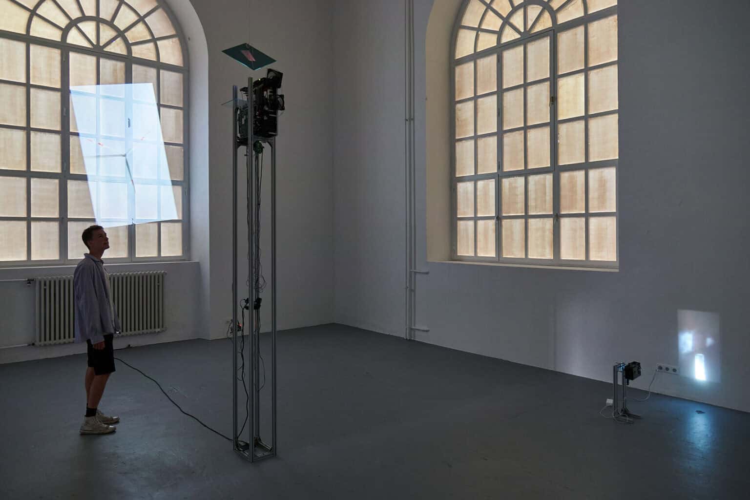 Foto: Ein abgedunkelter Raum mit zwei Fenstern. Im Raum stehen ein großes und ein kleines Gerüst, an denen Beamer angebracht sind. Die Beamer werfen auf unterschiedlichen Höhen Projektionen an die Wand. Eine der Projektionen ist nicht gerade sondern diagonal ausgerichtet und wird auf eines der Fenster mit abgedunkelten Scheiben projiziert.