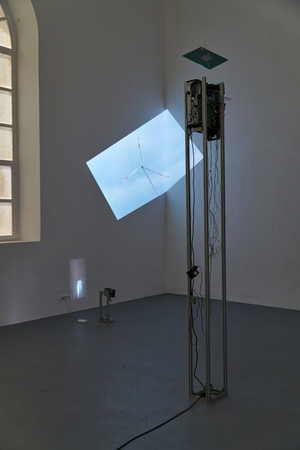 Foto: Ein abgedunkelter Raum. Im Raum stehen ein großes und ein kleines Gerüst, an denen Beamer angebracht sind. Die Beamer werfen auf unterschiedlichen Höhen Projektionen an die Wand. Eine der Projektionen ist nicht gerade sondern diagonal ausgerichtet und wird auf eine Raumecke projiziert.