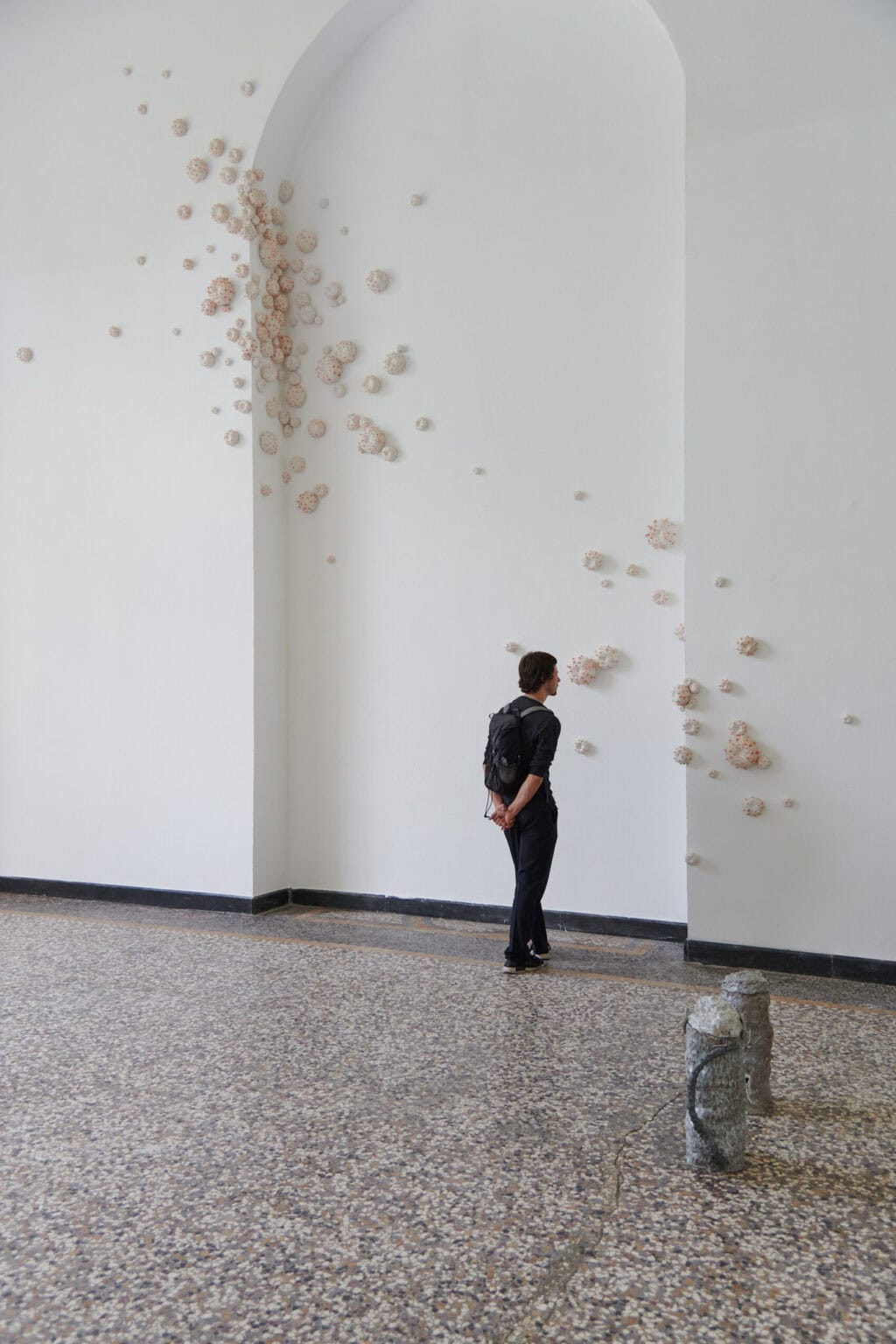 Foto: Ein Raum mit weißen Wänden, mehrfarbigem Steinboden und einer bogenförmigen Vertiefung in der Wand. Über die gesamte Höhe des Raumes verteilt befindet sich eine unregelmäßig angeordnete Gruppe von Objekten. Auf dem Boden steht ebenfalls ein Objekt, neben dem ein mensch steht.