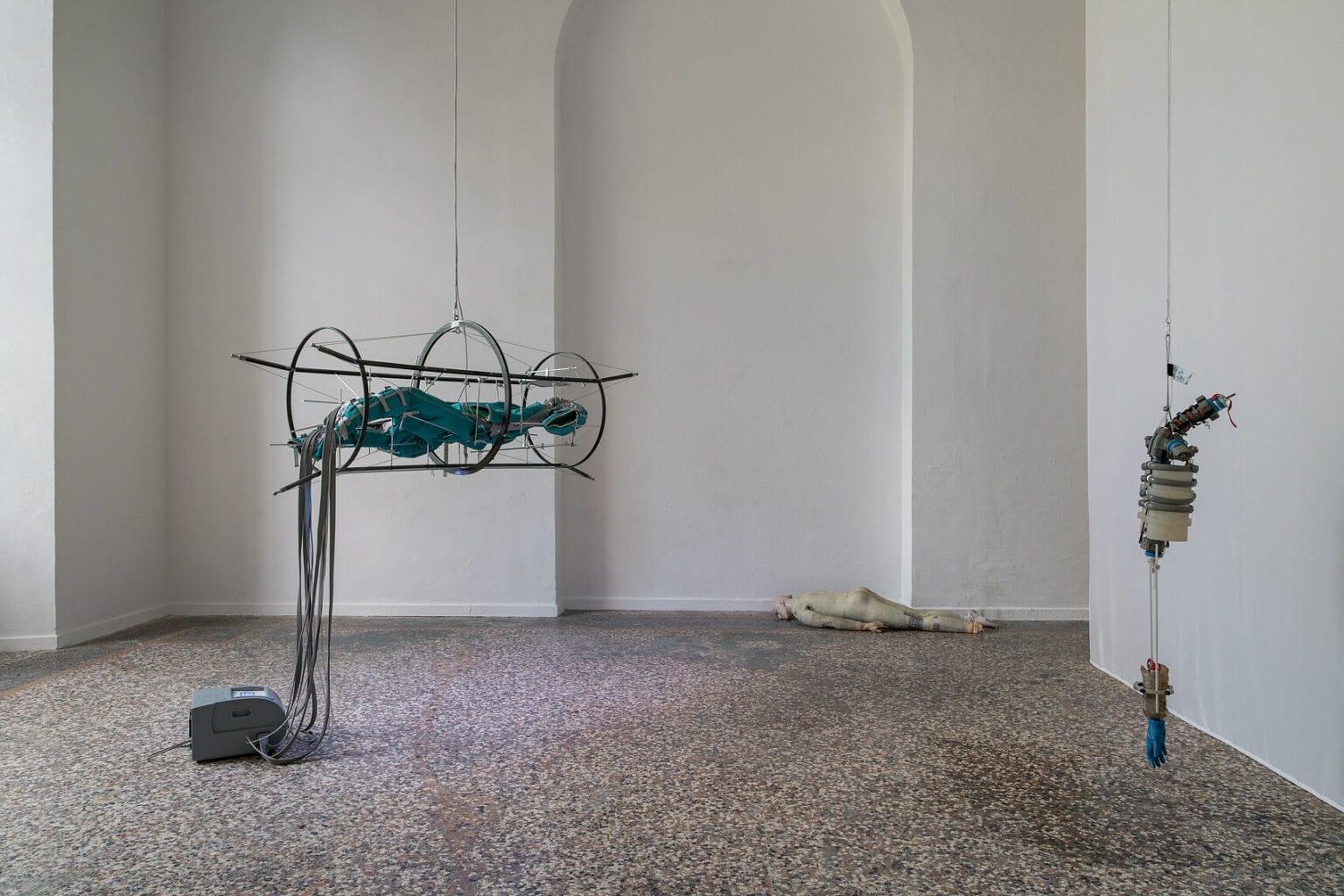 Foto: Ein Raum mit weißen Wänden und mehrfarbigem Steinboden. Im Raum verteilt befinden sich mehrere Objekte. Manche von ihnen hängen von der Decke, andere liegen auf dem Boden.