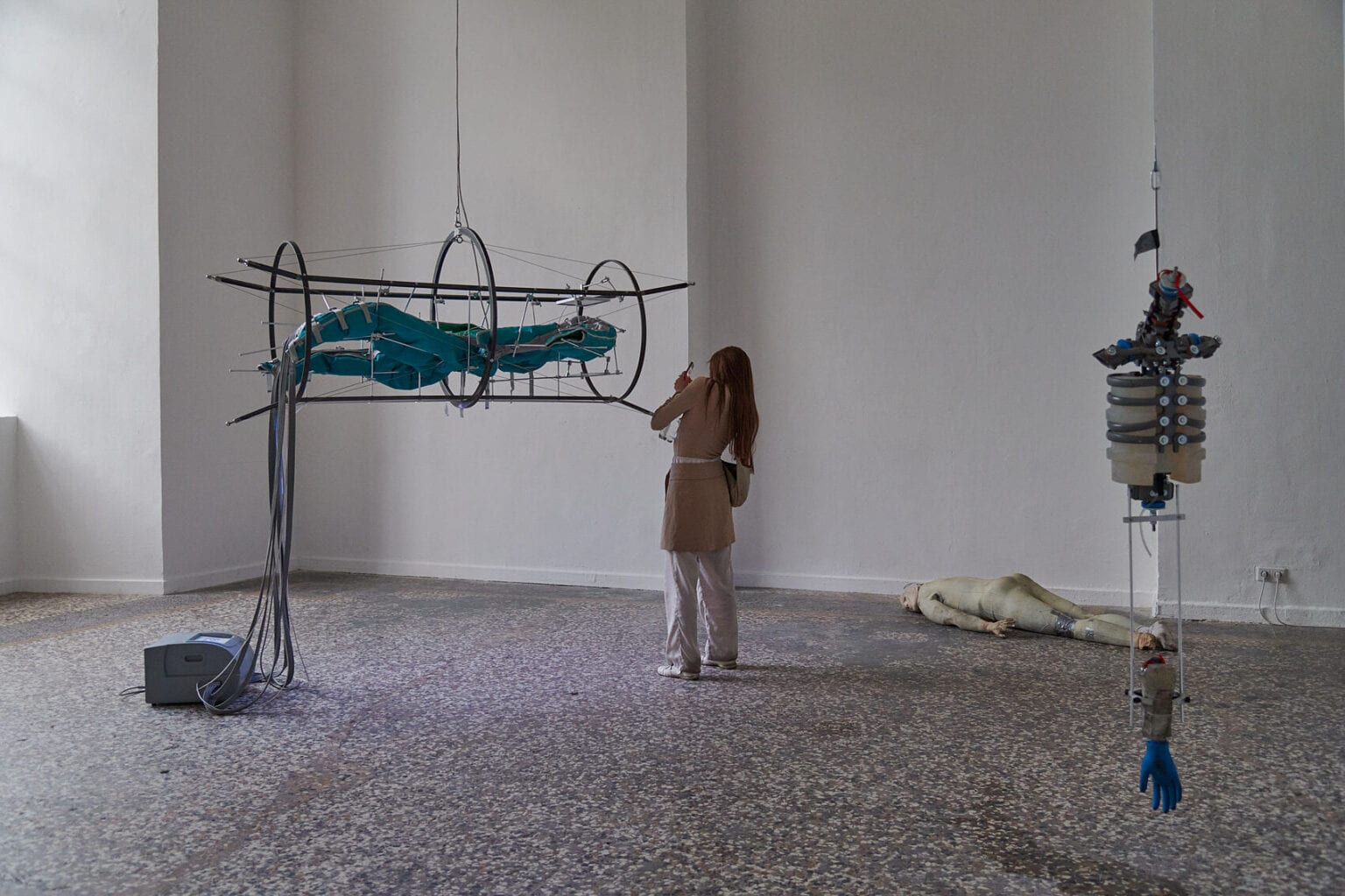 Foto: Ein Raum mit weißen Wänden und mehrfarbigem Steinboden. Im Raum verteilt befinden sich mehrere Objekte. Manche von ihnen hängen von der Decke, andere liegen auf dem Boden. Im Raum steht ein Mensch.