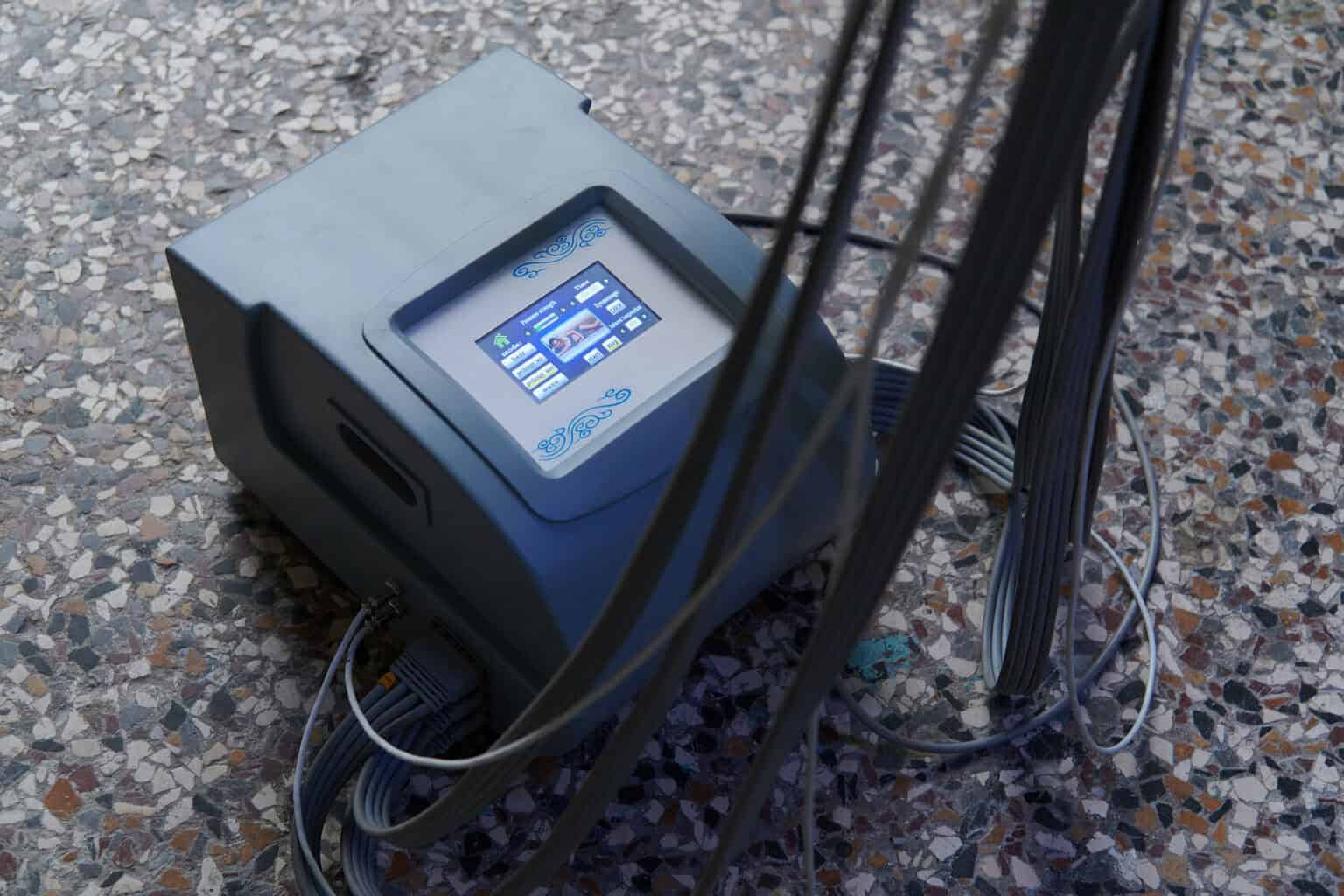 Foto: Ein Objekt mit Display von dem mehrere Kabel abgehen vor einem mehrfarbigen Steinboden.