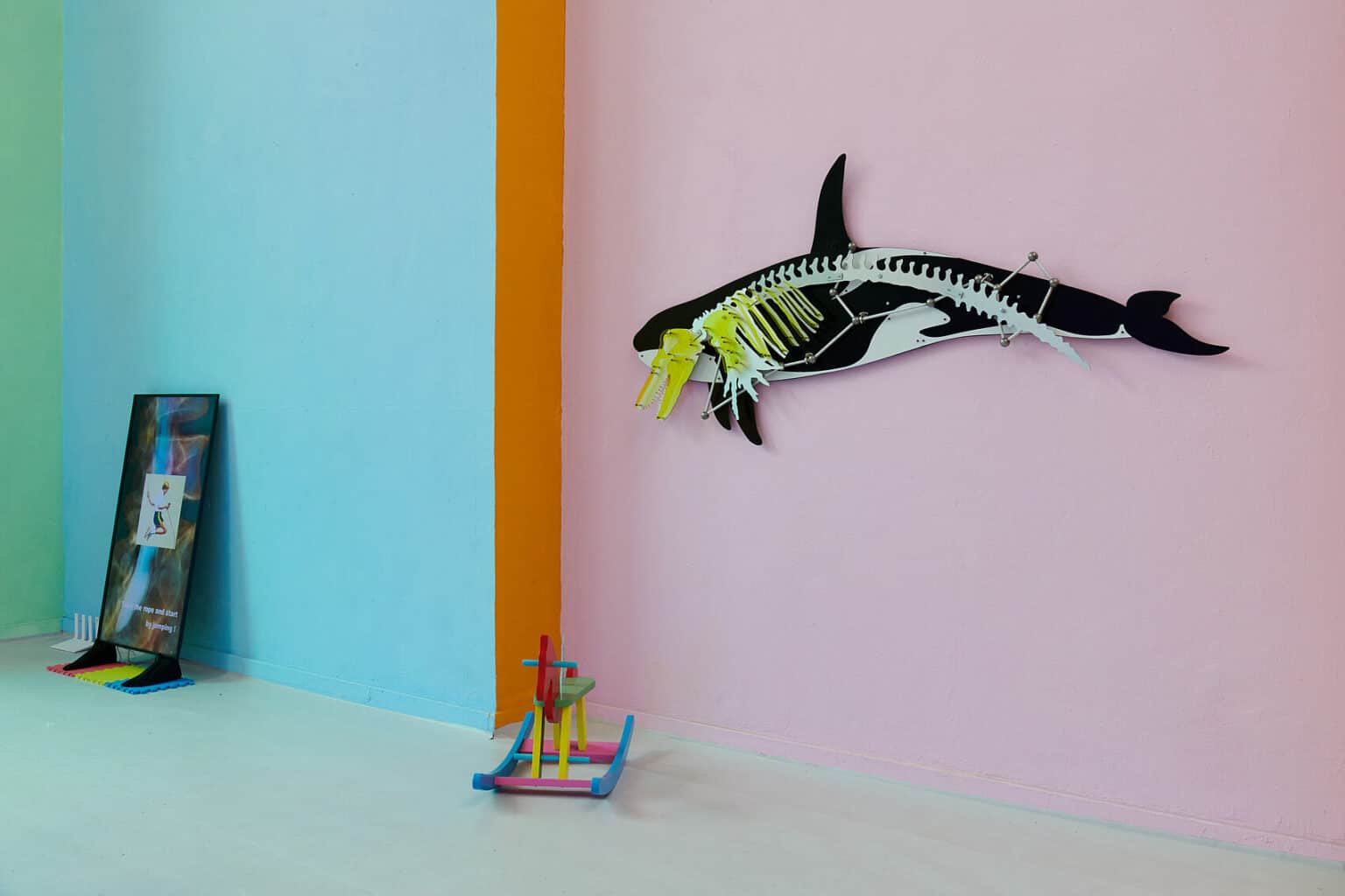 Foto: Ansicht eines Raumes mit bunt gestrichenen Wänden in den Farben grün, brau, orange und rosa. Der Boden ist grau. An der Wand, auf dem Boden Stehens und an der Wand lehnend befinden sich Objekte aus unterschiedlichen Materialien.