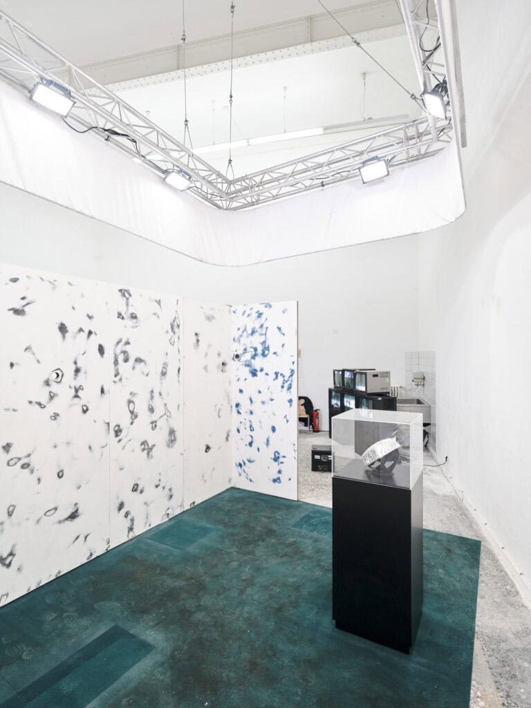 Foto: Ansicht eines Raumes mit weißen Wänden. Links im Bild befinden sich weiße Platten mit schwarzen, kreisrunden Abrieben. Der Boden ist mit einem grünen Material bedeckt, auf dem sich viele Schuhabdrücke abzeichnen. Auf dem grünen Material befindet sich ein schwarzer Sockel, auf dem ein Objekt und darüber ein transparenter Kasten sitzt. Im Hintergrund befindet sich ein Stapel aus Bildschirmen und ein Waschbecken aus Metall. An der Decke sind weiße Stahlträger zu sehen. Von der Decke hängt eine Konstruktion aus Metall und Textil, an die Lichter angebracht sind, die in den Raum leuchten.