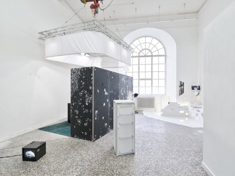 Foto: Ansicht eines Raumes mit weißen Wänden, mehrfarbigem Steinboden und einem großen Bogenfenster. Im Raum befinden sich eine Vielzahl von Objekten, darunter mehrere Bildschirme, weiße Backsteine, eine schwarze Wand mit weißen, kreisförmigen abreißen. Teile des Bodens sind mit weißem und grünen Material Bedeckt. Von der Decke hängt eine Konstruktion auf Metall und weißem Textil. in der Konstruktion befinden sich eingeschaltete Lichter.