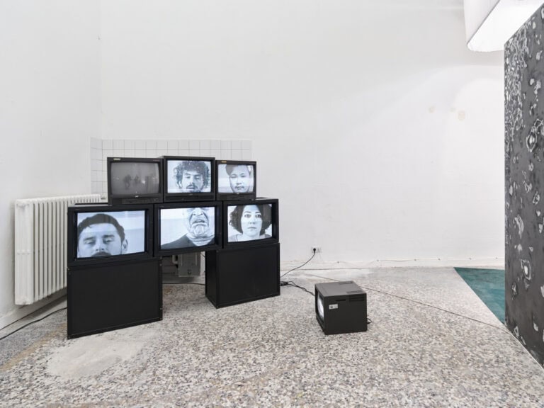 Foto: Ansicht eines Raumes mit weißen Wänden und mehrfarbigem Steinboden. Im Raum stehen Bildschirme, die aufeinander und auf schwarzen Kisten gestapelt sind.