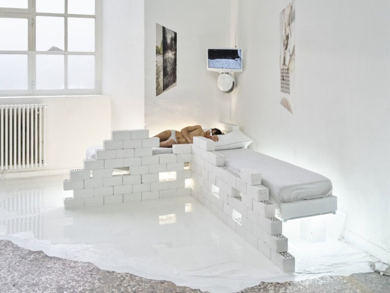 Foto: Aufnahme eines Raumes mit weißen Wänden und einem Fenster. Der Boden ist in großen Teilen mit einem weißen, reflektierenden Material bedeckt. In der Wecke des Raumes befindet sich eine Installation aus weißen Backsteinen und weiteren Materialien, darunter ein Bildschirm und flachen Objekten an der Wand, weißem Licht und Matratzen. Auf einer der Matratzen liegt ein Mensch, der eine weiße Unterhose und eine weiße Sonnenbrille trägt. Die Gläser der Brille sind mit weißer Farbe bedeckt.