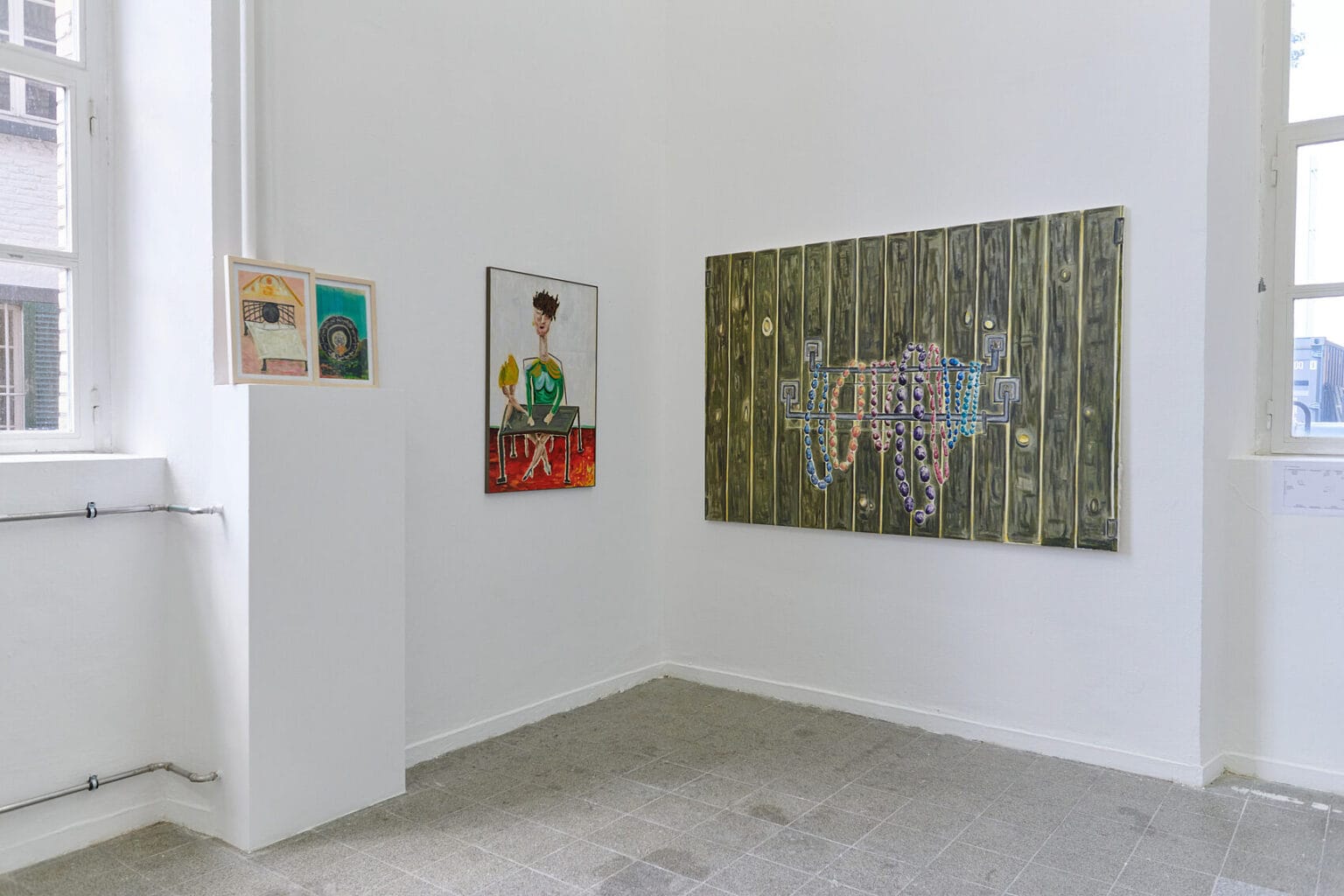 Foto: Die Ecke eines Raumes mit weißen Wänden und grauem, gefließten Boden. An den Wänden hängen, auf ähnlicher Höhe, flache Objekte im Klein-, Mittel- und Großformat.