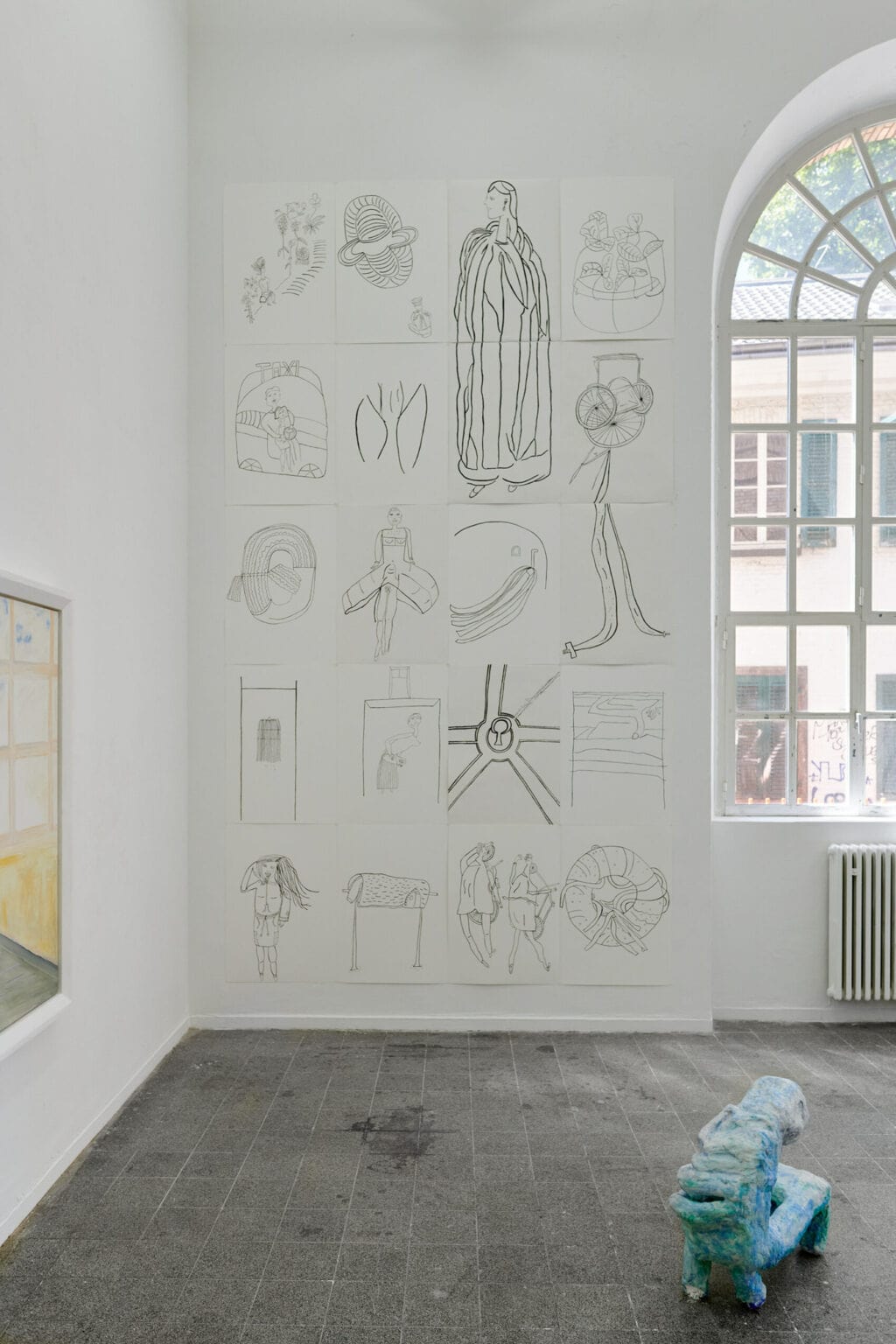 Foto: Ein Raum mit weißen Wänden, grauem, gefließten Boden und einem Fenster. An den Wänden eine Vielzahl flacher Objekte in unterschiedlichen Formaten und auf unterschiedlicher Höhe. Im Raum steht eine Skulptur auf dem Boden.