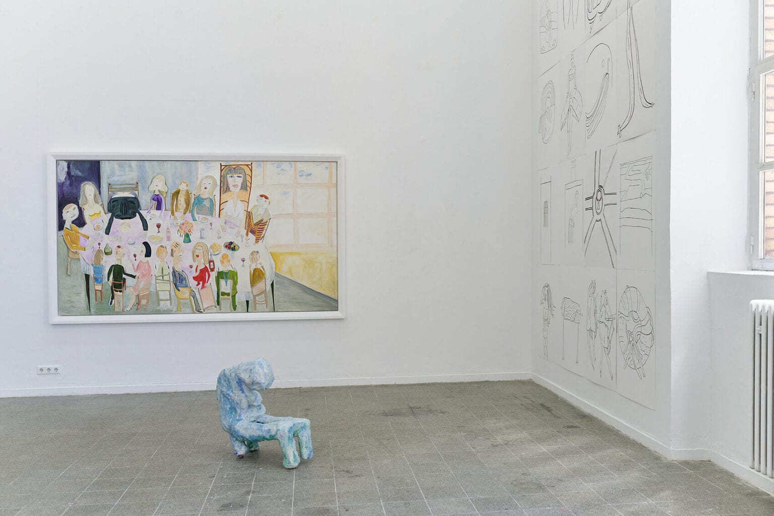 Foto: Ein Raum mit weißen Wänden, grauem, gefließten Boden und einem Fenster. An den Wänden eine Vielzahl flacher Objekte in unterschiedlichen Formaten und auf unterschiedlicher Höhe. Im Raum steht eine Skulptur auf dem Boden.