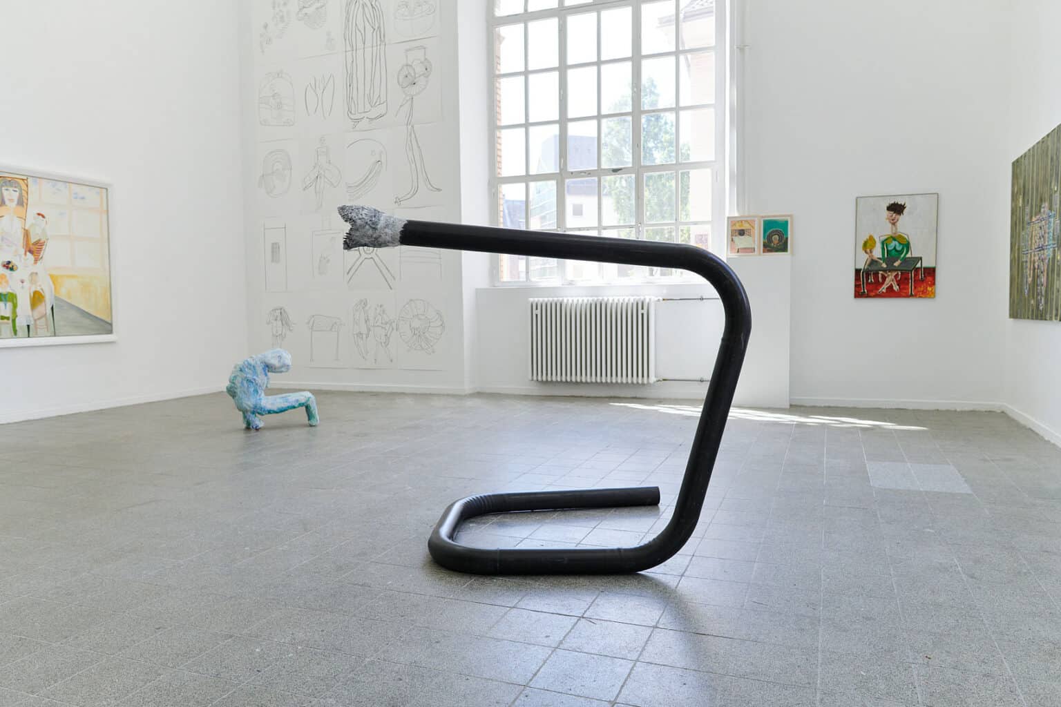 Foto: Ein Raum mit weißen Wänden, grauem, gefließten Boden und einem Fenster. An den Wänden eine Vielzahl flacher Objekte in unterschiedlichen Formaten und auf unterschiedlicher Höhe. Im Raum stehen zwei Skulpturen.