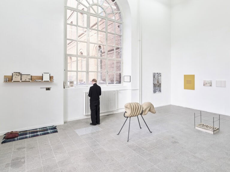 Foto: Ansicht eines Raumes mit weißen Wänden, grauem, gefliesten Boden und einem großen Bogenfenster. An den Wänden hängen Objekte. Auch auf dem Boden und auf der Fensterbank stehen mehrere Objekte. Vor dem Fenster steht eine Person, die zu Boden schaut.