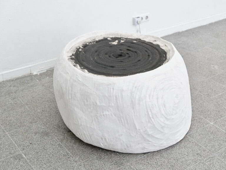 Foto: Nahaufnahme eines weißen, ründlichen Objektes vor einer weißen Wand und grauem, gefliesten Boden. Auf dem Objekt liegt eine schwarze, sandige Masse mit kreisförmigen Muster.