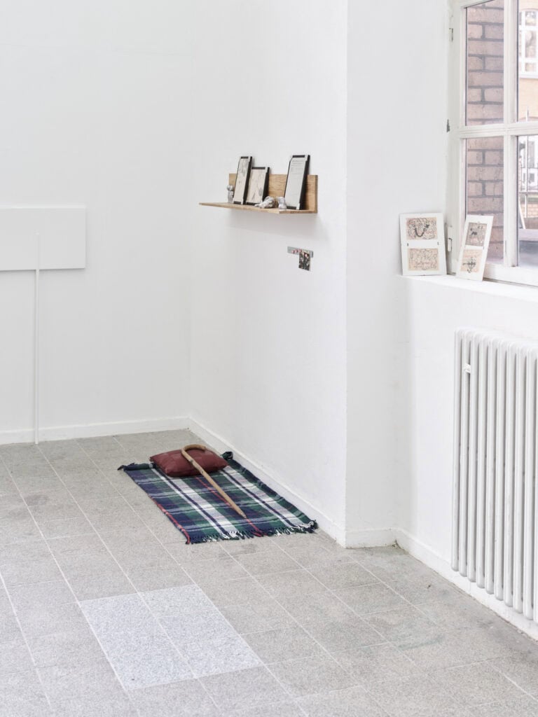 Foto: Ansicht einer Raumecke mit weißen Wänden und grauem, gefliesten Boden. An den Wänden hängen und auf dem Boden und der Fensterbank liegen Objekte.