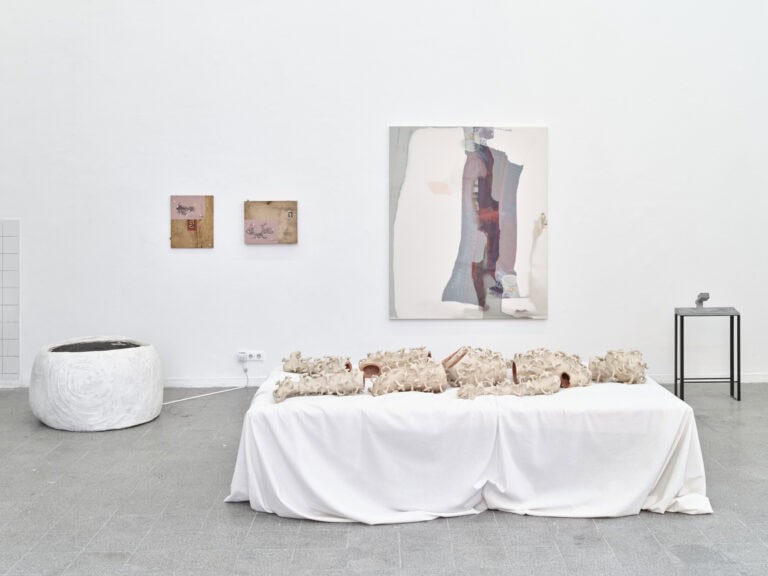 Foto: Ansicht eines Raumes mit weißen Wänden und grauem, gefliesten Boden. An den Wänden hängen klein- und großformatige Objekte. Auf dem Boden stehen mehrere Objekte.