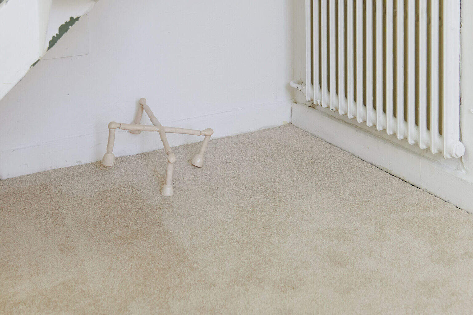 Foto: Ecke eines Raumes mit weißen Wänden und beigem Teppichboden. Auf dem Boden stehend und an die Wand gelehnt ein kleines, beiges Objekt.