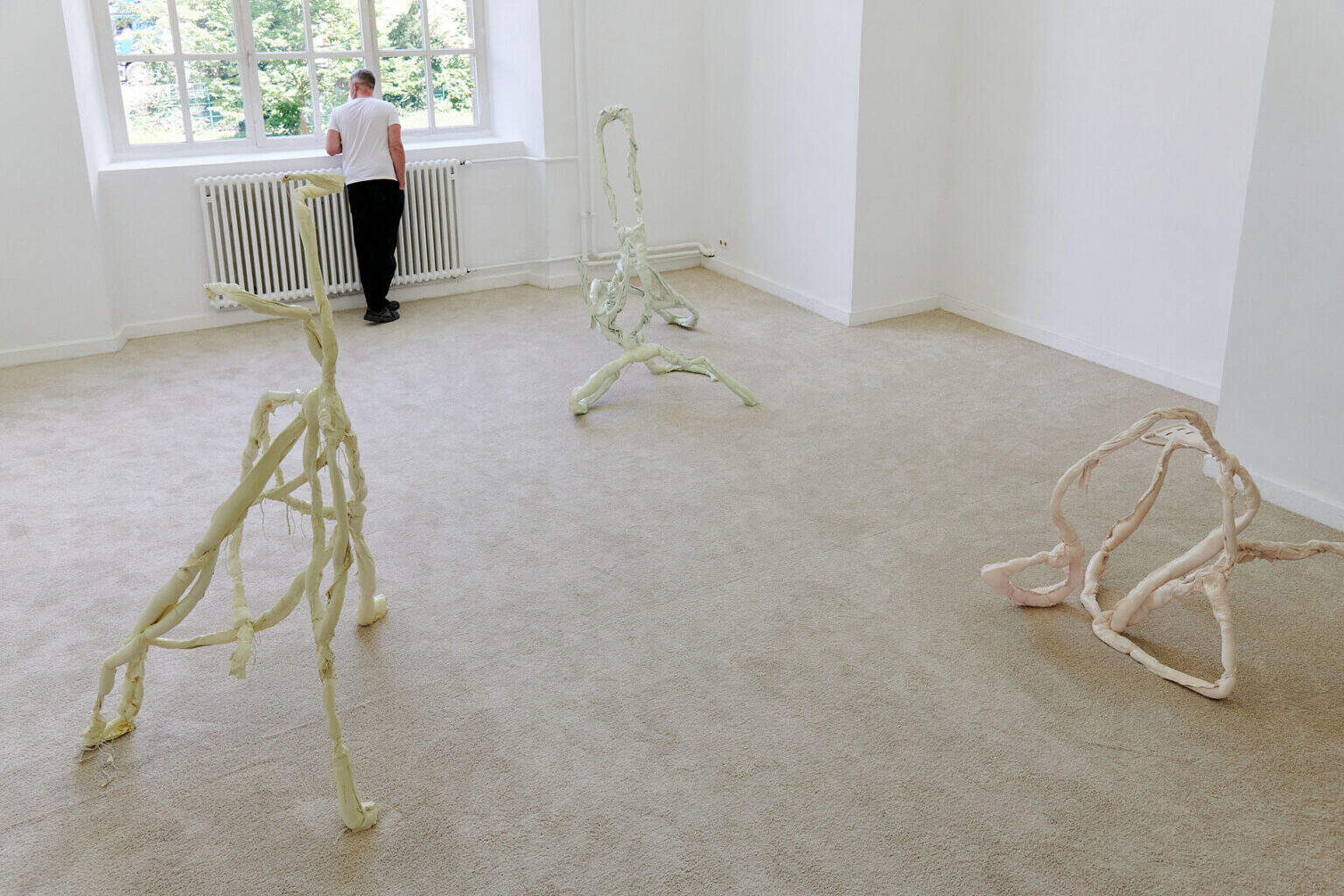 Foto: Ein Raum mit weißen Wänden, beigem Teppichboden und einem Fenster. Im Raum verteilt drei gewundene Skulpturen in Pastellfarben. Im Raum befindet sich eine Mensch.