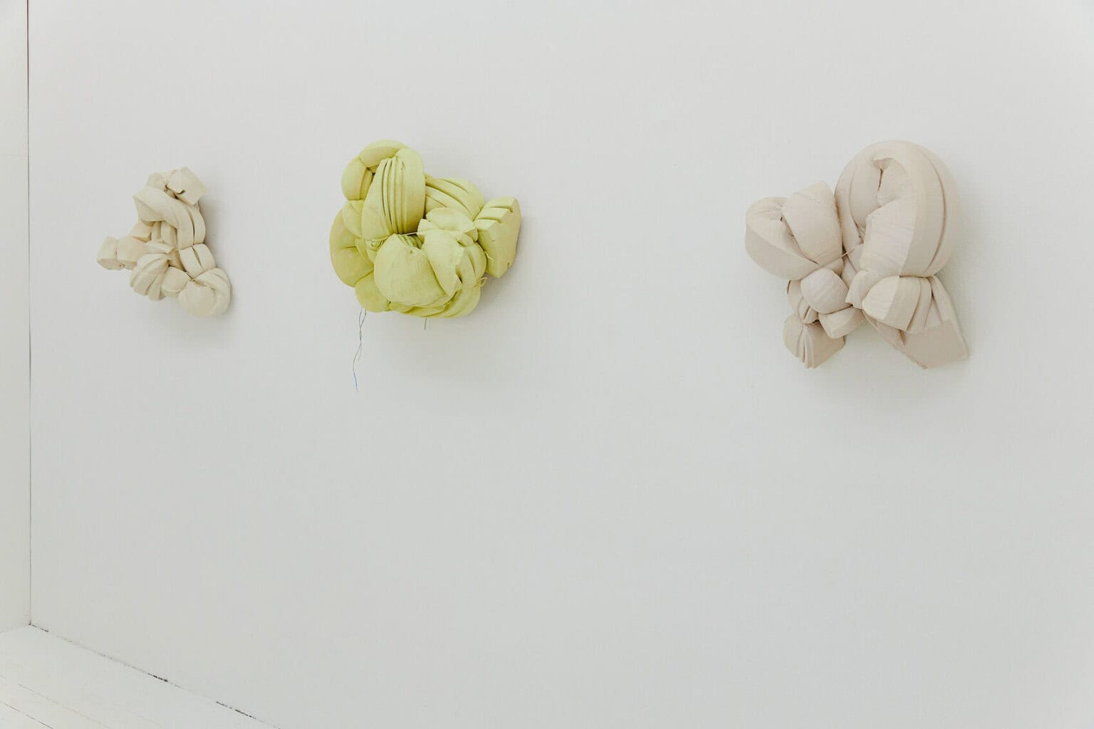 Foto: Drei in etwa gleich große, geschnürte Objekte, auf gleicher Linie hängend, vor weißer Wand.